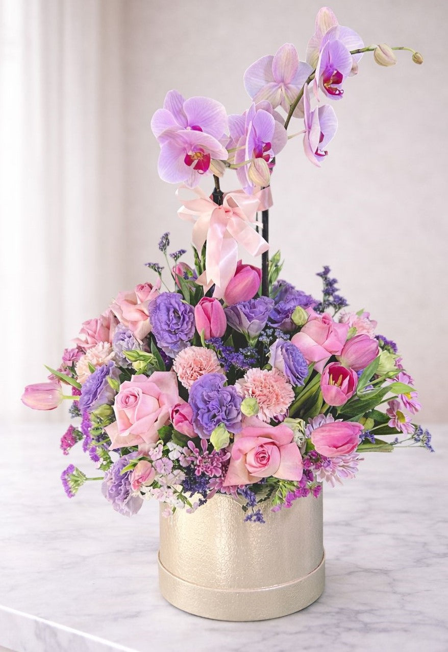 Arreglo floral premium con orquídea rosa, rosas, tulipanes y flores lilas en caja dorada elegante sobre mesa clara con luz natural, ideal para aniversario y regalo especial.