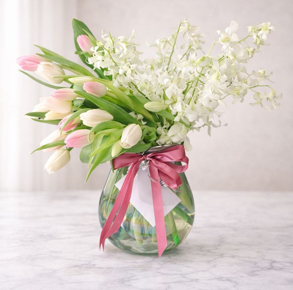 Arreglo floral elegante con tulipanes rosados y blancos y orquídeas blancas en jarrón de vidrio sobre mesa blanco marfil con luz natural, ideal para aniversario y regalo especial.