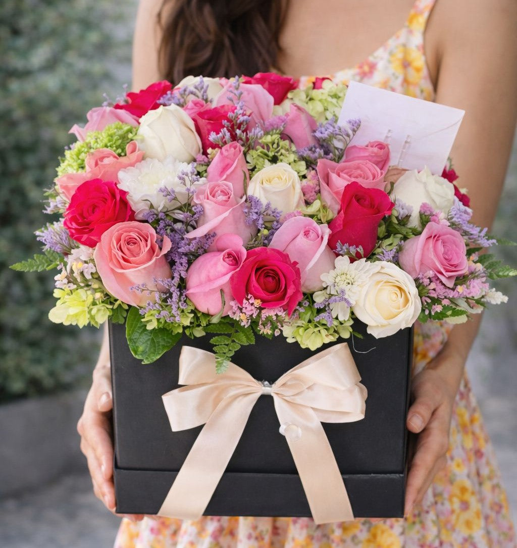 caja de flores con rosas en tonos rosa, blanco y fucsia sostenida por una mujer, arreglo floral elegante ideal para desear una pronta recuperación.